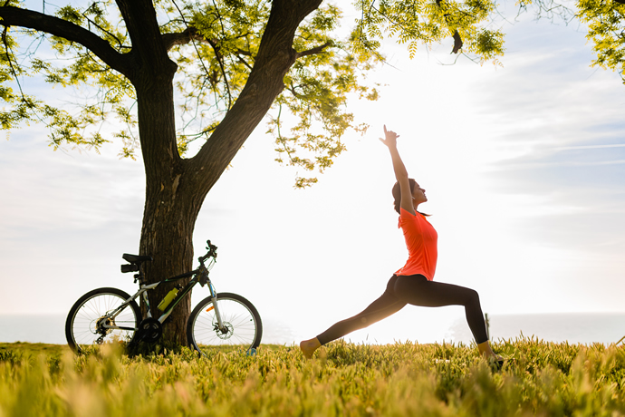 Kadın bir ağacın ve bisikletin yanında açık havada yoga yapıyor.